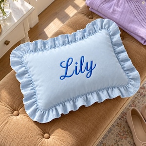 May include: A rectangular light blue throw pillow featuring a ruffled border and the name Lily embroidered in dark blue script lettering centred on the front. The pillow is constructed with a soft fabric finish and a consistent ruffled edge.