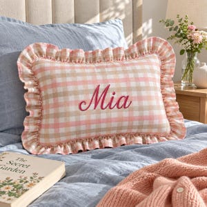 May include: A rectangular throw pillow featuring a pink and beige gingham check pattern with a ruffled border. The name Mia is embroidered in dark pink cursive script across the centre of the pillow. The pillow is made of fabric.