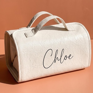 Puede incluir: Un bolso de viaje rectangular de color blanco roto con una superficie texturizada y arrugada, y dos asas superiores. El nombre Chloe está impreso en cursiva negra en el panel frontal del bolso, que presenta un diseño estructurado.