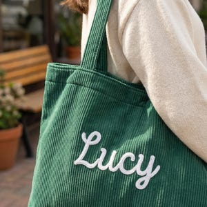 以下が含まれることがあります： 緑色のコーデュロイのトートバッグ。白い筆記体で「Lucy」と刺繍されています。バッグは長方形で、幅広のショルダーストラップが付いています。背景には、クリーム色のセーターを着た人が写っています。