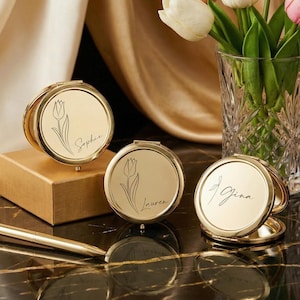 Op de afbeelding: Drie goudkleurige, ronde compacte spiegels met gepersonaliseerde namen en bloemmotieven. De namen "Sophie", "Lauren" en "Gina" zijn sierlijk geschreven. Een gouden pen ligt op een donker, patroon oppervlak, met een vaas met witte tulpen op de achtergrond.