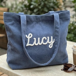 Puede incluir: Un bolso tote de pana azul con el nombre "Lucy" bordado en escritura blanca. El bolso tiene dos asas y forma rectangular. Gafas de sol y una taza de café en el fondo.