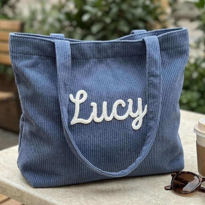 以下が含まれることがあります： 白い筆記体で「Lucy」と刺繍された青いコーデュロイのトートバッグ。 バッグには2つのハンドルと長方形の形があります。 サングラスとコーヒーカップが背景にあります。