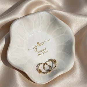 K&ouml;nnte beinhalten: Eine wei&szlig;e, blumenf&ouml;rmige Keramikschale mit gl&auml;nzender Oberfl&auml;che und dezenter Blattaderstruktur. In der Mitte befindet sich eine goldene Aufschrift j & c Engaged May.20.26. Zwei goldene Creolen liegen in der Schale.