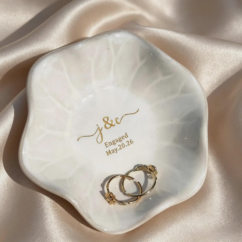 K&ouml;nnte beinhalten: Eine wei&szlig;e, blumenf&ouml;rmige Keramikschale mit gl&auml;nzender Oberfl&auml;che und dezenter Blattaderstruktur. In der Mitte befindet sich eine goldene Aufschrift j & c Engaged May.20.26. Zwei goldene Creolen liegen in der Schale.