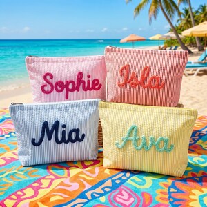 Puede incluir: Cuatro neceseres personalizados con nombres bordados. Las bolsas son rosas, rojas, azules y amarillas con rayas verticales. Los nombres son Sophie, Isla, Mia y Ava. Las bolsas están sobre una toalla de playa colorida con una playa y un océano de fondo.