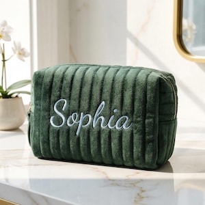 May include: A green, rectangular wash bag with a ribbed texture. The name "Sophia" is embroidered in white script on the front. The bag has a gold zip and is likely made of a soft, plush material.