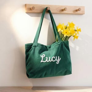 以下が含まれることがあります： 白い筆記体で「Lucy」と刺繍された緑色のコーデュロイのトートバッグ。バッグは3つのペグが付いた木製のラックに吊り下げられており、黄色の水仙で満たされています。バッグには長いハンドルが付いています。