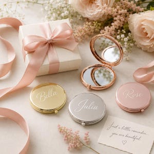 Op de afbeelding: Drie ronde metalen compacte spiegels in goud, zilver en roségoud, elk gegraveerd met de namen Bella, Julia en Rose. Eén spiegel is geopend en toont twee reflecterende oppervlakken. Een geschenkdoos met een roze lint en een briefje zijn inbegrepen.
