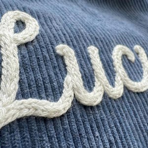 以下が含まれることがあります： 青色のコーデュロイ生地のクローズアップ。白い筆記体で「Lucy」という名前が刺繍されています。白い刺繍は厚く、質感のある外観で、青い生地の柔らかくリブのある質感と対照的です。