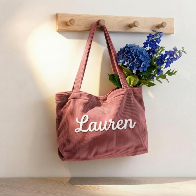 以下が含まれることがあります： 「Lauren」の白い文字が刺繍された、くすんだピンク色のコーデュロイのトートバッグ。バッグには青い花が詰められ、木製のペグラックに掛けられています。バッグには2つの長いハンドルが付いています。