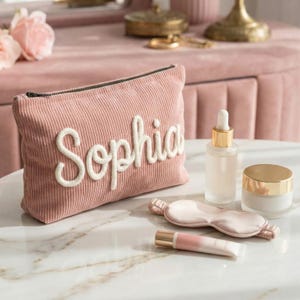 Puede incluir: Un neceser de pana rosa con el nombre "Sophia" bordado en letras blancas. Junto a él hay un frasco con cuentagotas, un tarro con tapa dorada, una máscara para dormir y un tubo de brillo labial.