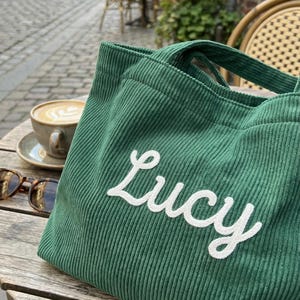 以下が含まれることがあります： 白い筆記体で「Lucy」と刺繍された緑色のコーデュロイのトートバッグ。バッグは、コーヒーカップとサングラスの隣の木製のテーブルの上にあります。背景にはヨーロッパの街並みが写っています。