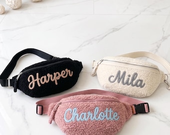 Puede incluir: Tres riñoneras de tela peluda con correas ajustables y cierres de cremallera. El bolso negro lleva el nombre Harper en color melocotón, el crema muestra el nombre Mila en gris y el rosa presenta el nombre Charlotte en azul claro.