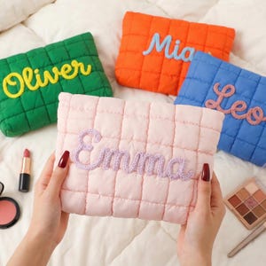 Op de afbeelding: Een verzameling gewatteerde make-up tasjes in groen, oranje, blauw en roze. Elke tas heeft een naam geborduurd in een contrasterende kleur: Oliver, Mia, Leo en Emma. De rechthoekige tassen hebben een rasterpatroon.