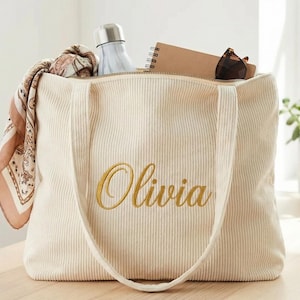 Puede incluir: Bolso tote de pana color crema con el nombre "Olivia" bordado en oro. El bolso abierto revela una bufanda estampada, una botella de agua, un cuaderno y gafas de sol. El bolso tiene asas largas.