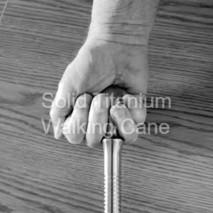 May include: A grayscale image of a solid titanium walking cane. The cane is held in a hand, with the text "Solid Titanium Walking Cane" visible. The cane has a ribbed design.
