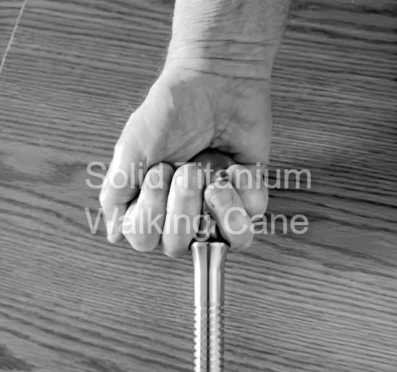 May include: A grayscale image of a solid titanium walking cane. The cane is held in a hand, with the text "Solid Titanium Walking Cane" visible. The cane has a ribbed design.
