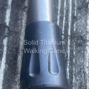 May include: Close-up of a solid titanium walking cane. The image shows the black rubber tip and the silver metal shaft. The text "Solid Titanium Walking Cane" is printed on the cane.