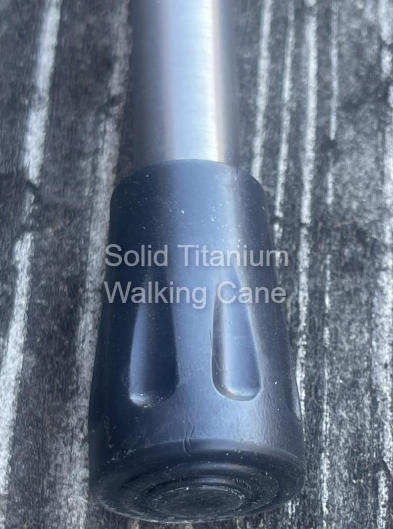 May include: Close-up of a solid titanium walking cane. The image shows the black rubber tip and the silver metal shaft. The text "Solid Titanium Walking Cane" is printed on the cane.