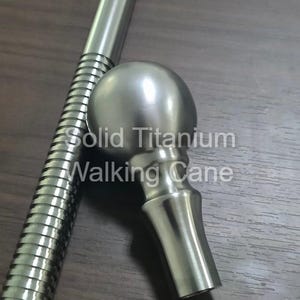 May include: A solid titanium walking cane with a rounded handle and a ribbed shaft. The text "Solid Titanium Walking Cane" is visible on the image. The cane is silver in color and appears to be made of metal.