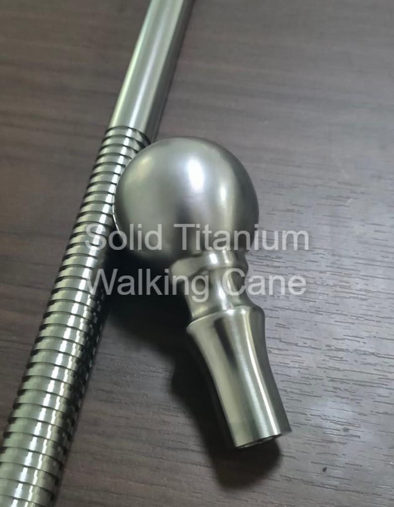 May include: A solid titanium walking cane with a rounded handle and a ribbed shaft. The text "Solid Titanium Walking Cane" is visible on the image. The cane is silver in color and appears to be made of metal.