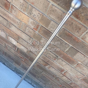 May include: A silver-colored walking cane with a rounded top and a textured grip section. The text "Solid Titanium Walking Cane" is printed on the brick background. The cane is angled against a brick wall.