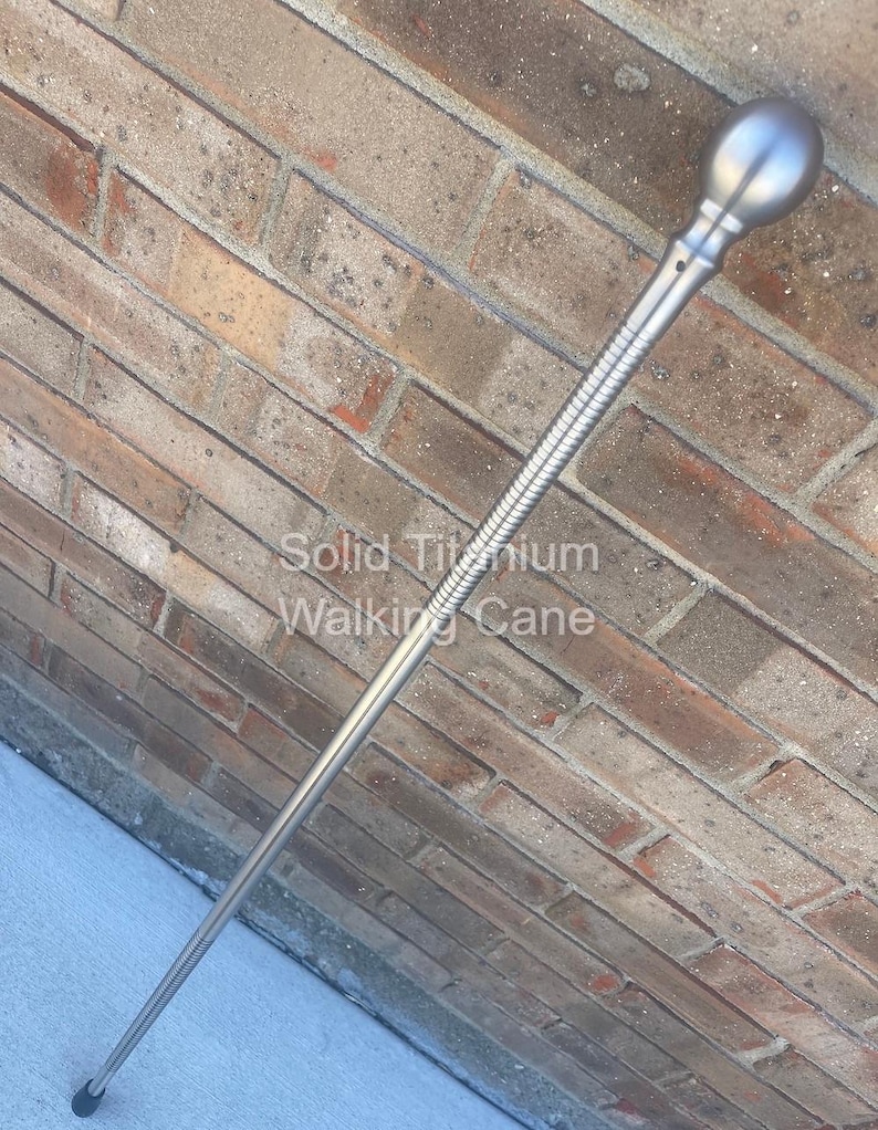 May include: A silver-colored walking cane with a rounded top and a textured grip section. The text "Solid Titanium Walking Cane" is printed on the brick background. The cane is angled against a brick wall.