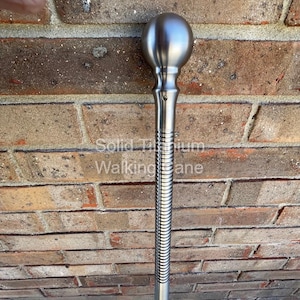 May include: A silver-colored walking cane with a spherical handle. The shaft has a ribbed design, and the text "Solid Titanium Walking Cane" is visible. The cane is set against a brick wall.