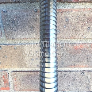 May include: A close-up of a metallic silver walking cane with a ridged design. The cane is set against a weathered brick wall. The text "Solid Titanium Walking Cane" is visible on the cane.