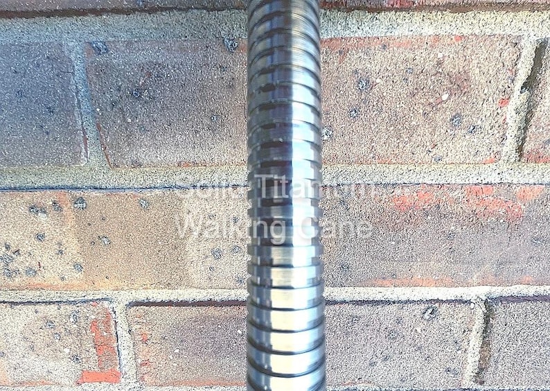 May include: A close-up of a metallic silver walking cane with a ridged design. The cane is set against a weathered brick wall. The text "Solid Titanium Walking Cane" is visible on the cane.