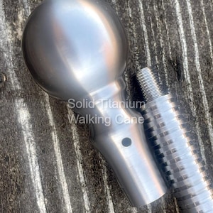 May include: A close-up of a solid titanium walking cane. The cane features a rounded handle and a textured shaft. The text "Solid Titanium Walking Cane" is visible on the image. The cane is silver in color.