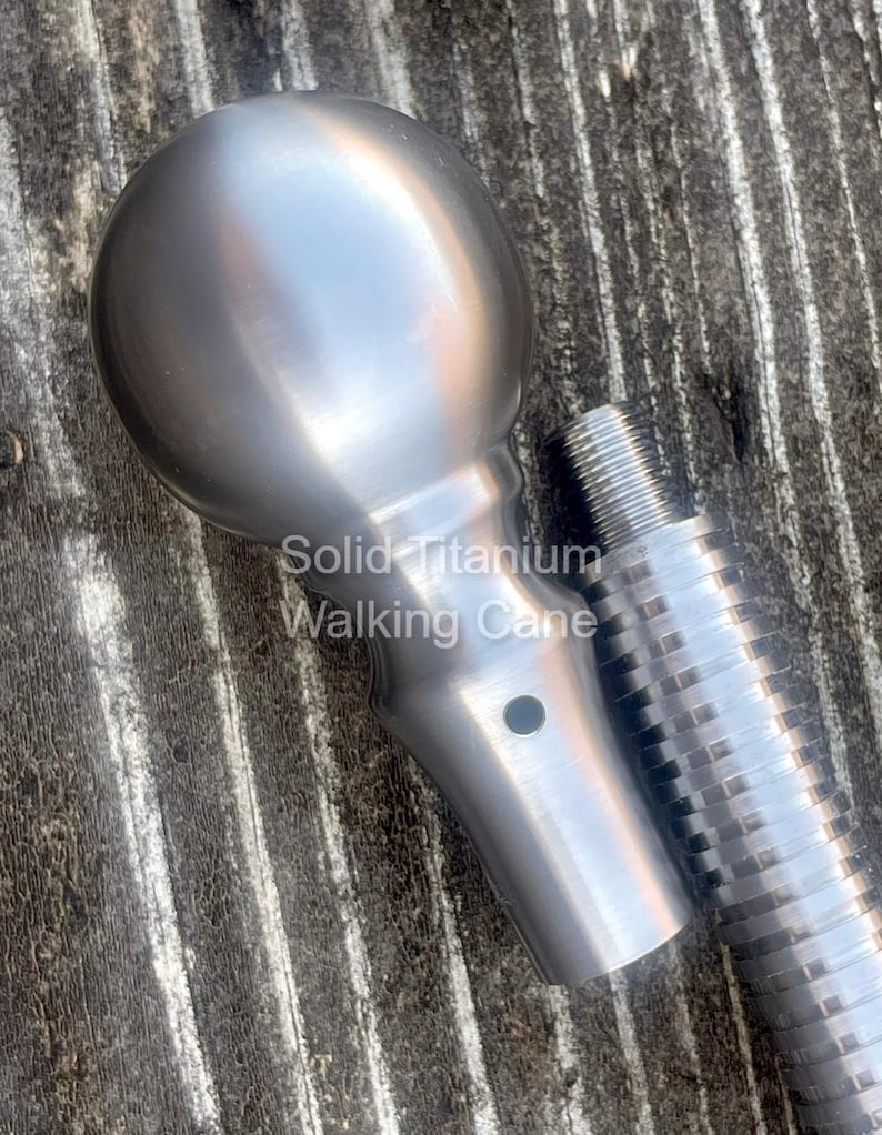 May include: A close-up of a solid titanium walking cane. The cane features a rounded handle and a textured shaft. The text "Solid Titanium Walking Cane" is visible on the image. The cane is silver in color.