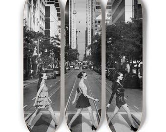 May include: Set of three skateboard decks featuring a black and white image of women crossing a city street. The decks are oval-shaped with a cityscape background. The women are walking on a crosswalk.