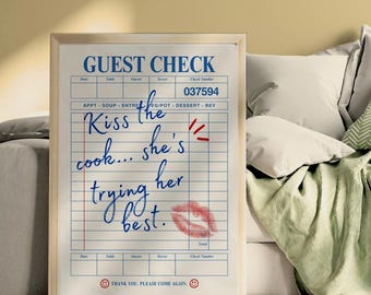 May include: A framed print of a guest check with the words "Kiss the cook... she's trying her best" written in blue ink. A red lipstick kiss mark is on the check. The check has a wooden frame.