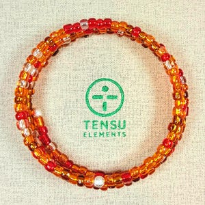 May include: A multi-strand wrap bracelet composed of small, translucent glass beads in shades of orange, red, and yellow. The circular bracelet surrounds a green logo featuring a crosshair symbol and the text TENSU ELEMENTS.