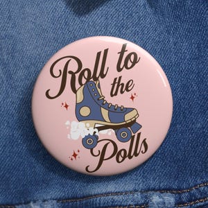 May include: A round pin-back button with a light pink background featuring a dark blue roller skate illustration, white motion clouds, small red star accents, and the dark brown text "Roll to the Polls" written in a cursive script font.