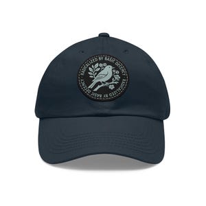 May include: A navy blue baseball cap featuring a circular black patch on the front. The patch displays a light blue illustration of a bird perched on a flowering branch, surrounded by the text "RADICALIZED BY BASIC DECENCY" in a circular font.