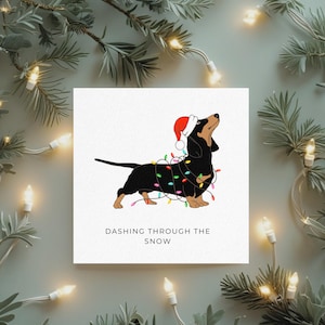 May include: A Christmas card featuring a black and tan dachshund wearing a Santa hat and wrapped in colorful Christmas lights. The text "Dashing Through The Snow" is below the dog on a white background.