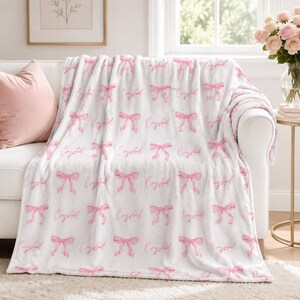 Peut inclure: Un plaid blanc en peluche orné d'un motif répétitif de nœuds rose tendre et du prénom Crystal écrit en lettres cursives. La couverture présente une texture duveteuse et est drapée sur un canapé blanc, mettant en valeur les tons rose et blanc.