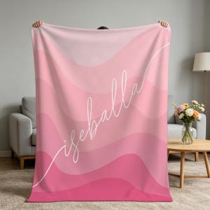 May include: A rectangular throw blanket featuring a gradient of pink wavy horizontal stripes. The name "isabella" is printed in a white, cursive, script-style font diagonally across the center of the blanket, extending from the bottom left corner.