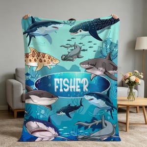 Peut inclure: Une couverture en polaire bleu clair ornée d'un motif de dessin animé représentant diverses espèces de requins, dont des requins-marteaux et des requins-baleines, nageant sous l'eau. Un ovale bleu foncé au centre porte le nom FISHER en blanc.
