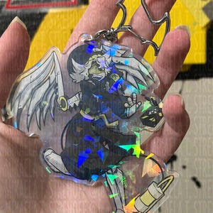 May include: A clear acrylic keychain featuring a character with white wings, a sword, and a dark outfit. The keychain has a silver-colored bat-shaped clasp. The design incorporates holographic elements, creating a shimmering effect.