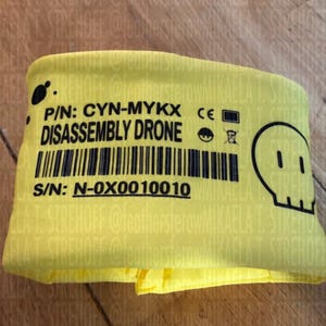 Puede incluir: Un artículo de tela amarilla doblada con texto negro y un código de barras. El texto dice "DISASSEMBLY DRONE" e incluye un número de producto y de serie. Un gráfico de calavera negra está en el lado derecho.