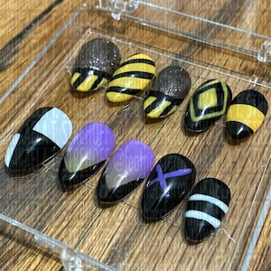 Könnte beinhalten: Ein Set aus zehn künstlichen Nägeln in einer transparenten Kunststoffbox. Die Nägel weisen verschiedene Designs auf, darunter Schwarz-Weiß, gelb-schwarze Streifen, Glitzer und lila Ombre. Die Nägel sind oval geformt.