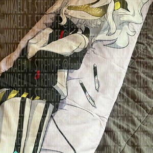 Op de afbeelding: Een lange rechthoekige kussensloop met een print van een anime-figuur met wit haar, gekleed in een zwart-wit gestreepte outfit met gele accenten. Het ontwerp bevat twee zwart-witte pennen die op de stof liggen.