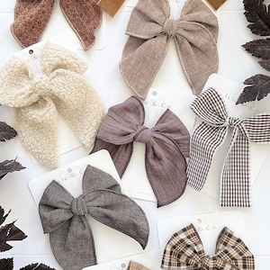 May include: A collection of hair bows in various textures and patterns, including brown floral, tan velvet, beige sherpa, solid brown linen, dark brown muslin, brown gingham, dark gray linen, tan plaid, and light brown embroidered fabric on display cards.