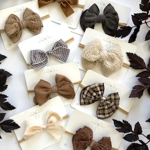 May include: A collection of eight hair bows on individual white display cards labeled Ivie & Co. The bows feature various textures and patterns, including brown velvet, charcoal linen, black and white gingham, cream sherpa, brown corduroy, brown plaid, cream dotted fabric, and brown floral print.
