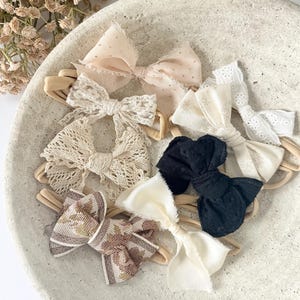 Puede incluir: Una colección de seis lazos para el cabello sujetos a finas bandas elásticas de colores neutros. Los lazos presentan diversas texturas y patrones, incluyendo tul beige con lunares, encaje blanco con ojales, encaje crema, tela negra y un estampado floral marrón.