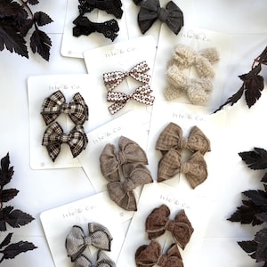 May include: A collection of hair bows in various textures and patterns, including black lace, charcoal gray, cream sherpa, brown plaid, tan linen, and striped fabric. Each pair is mounted on a white Ivie & Co. branded display card.